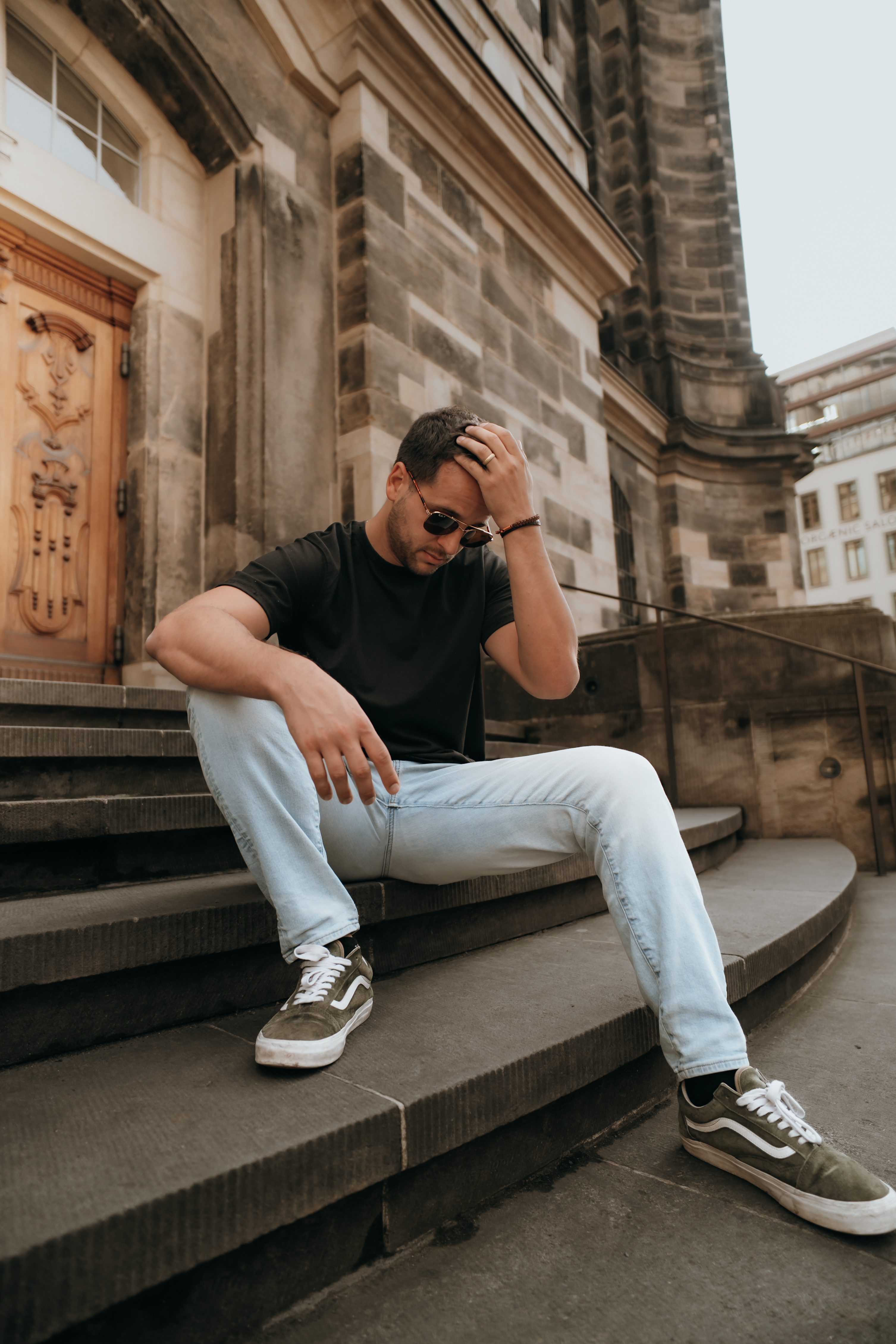 A man sitting on stone steps, wearing a black t-shirt and light blue jeans, with sunglasses and a contemplative expression.