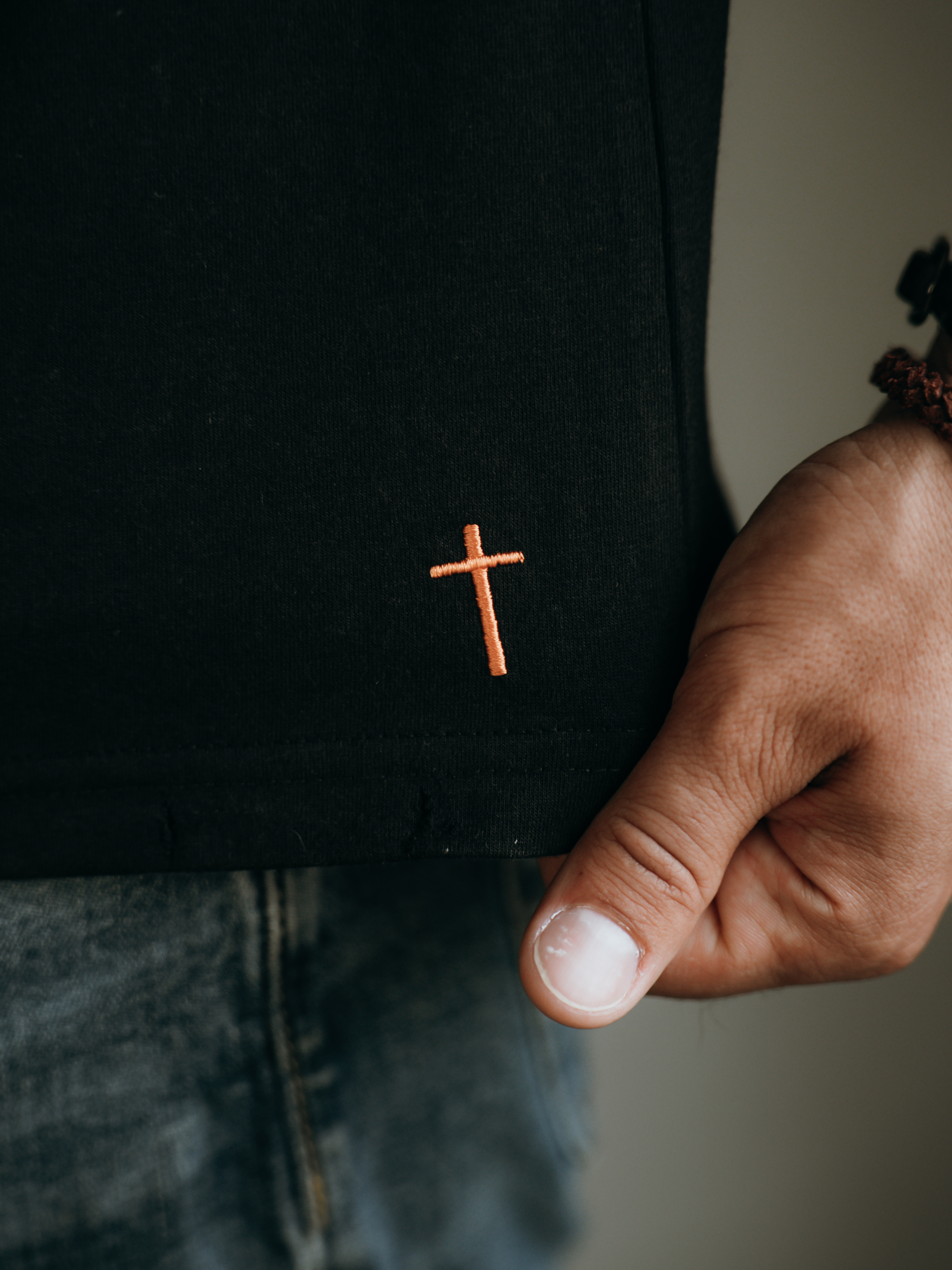 A close-up of a person's hand holding the hem of a black t-shirt with an orange cross embroidered on it.